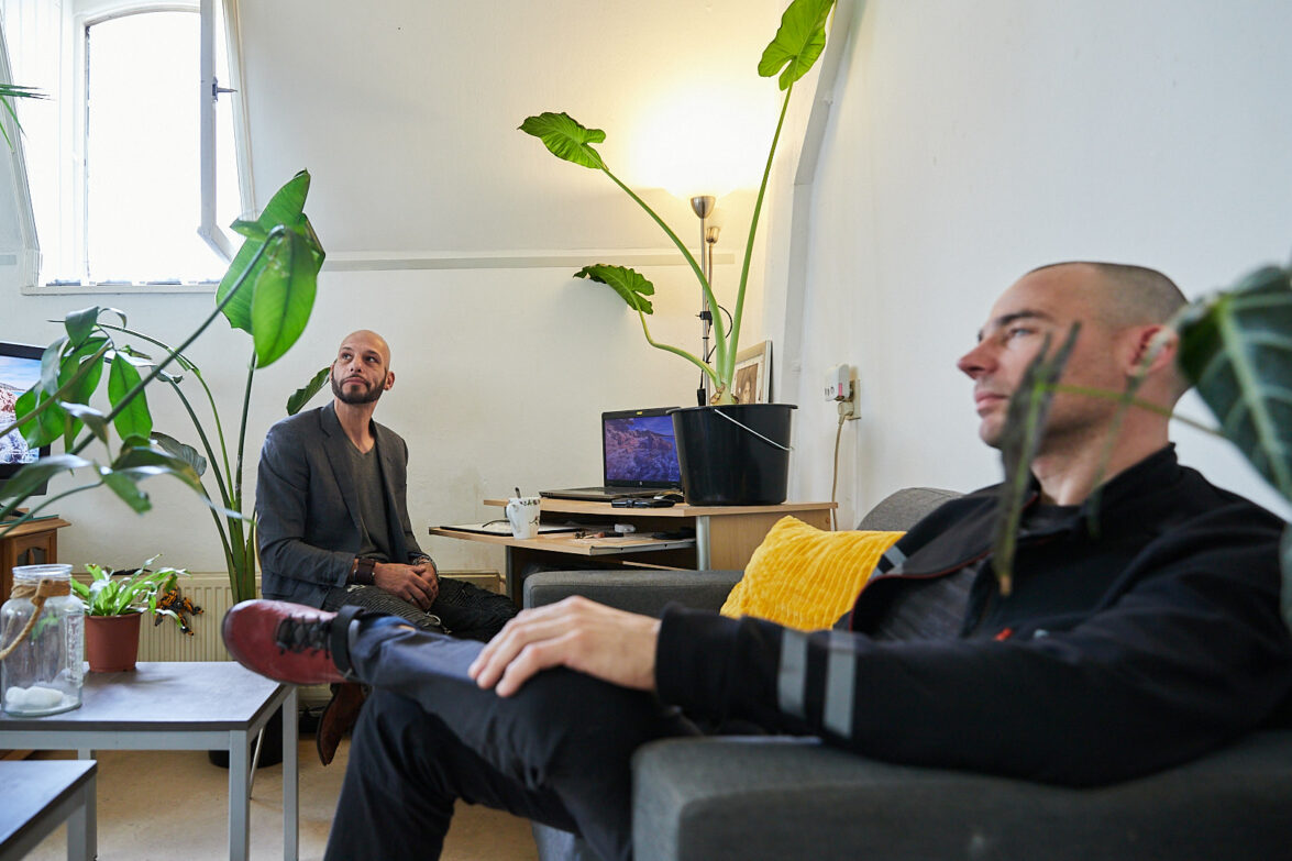 Twee mannen (cliënt en persoonlijk coach) zitten samen in de studio van de cliënt en kijken naar een persoon buiten beeld.